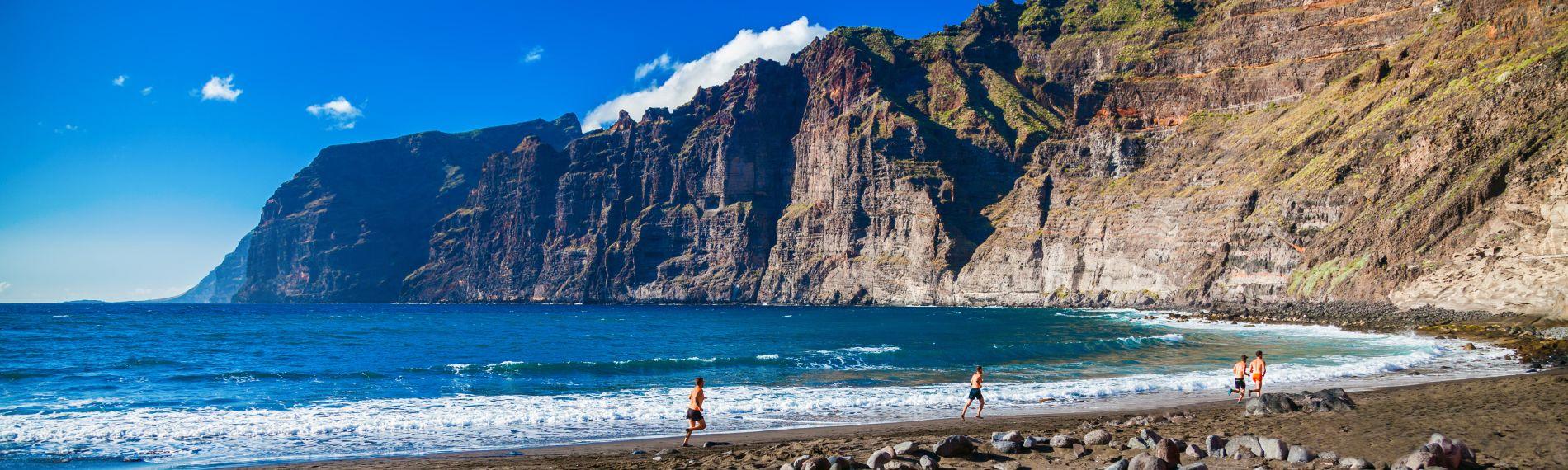 Four runners following the waterline on Playa de los Guios beach, where Tenerife's famous Los Gigantes cliffs rise up beside the black sands and blue sea near Puerto de Santiago