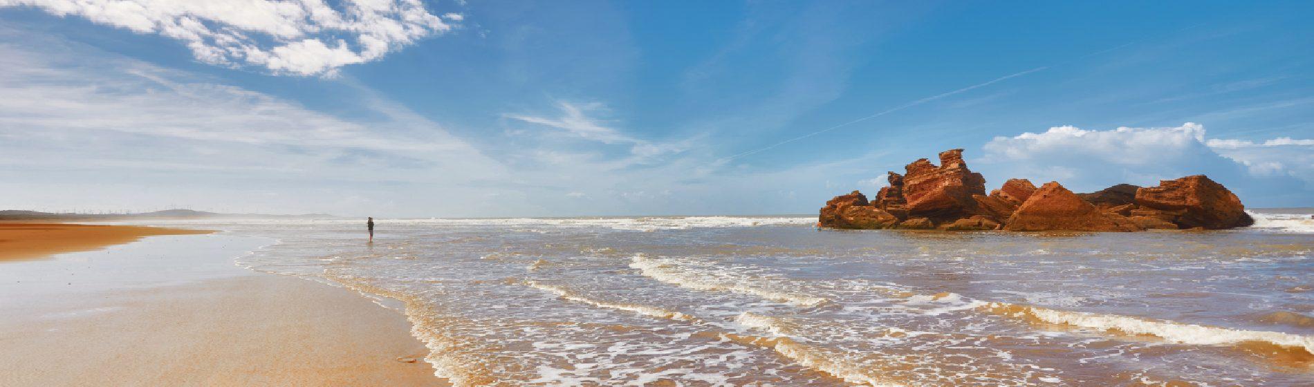 Sandy beach in Morocco with foamy waves, red rock formations, and a lone walker under a bright blue sky.