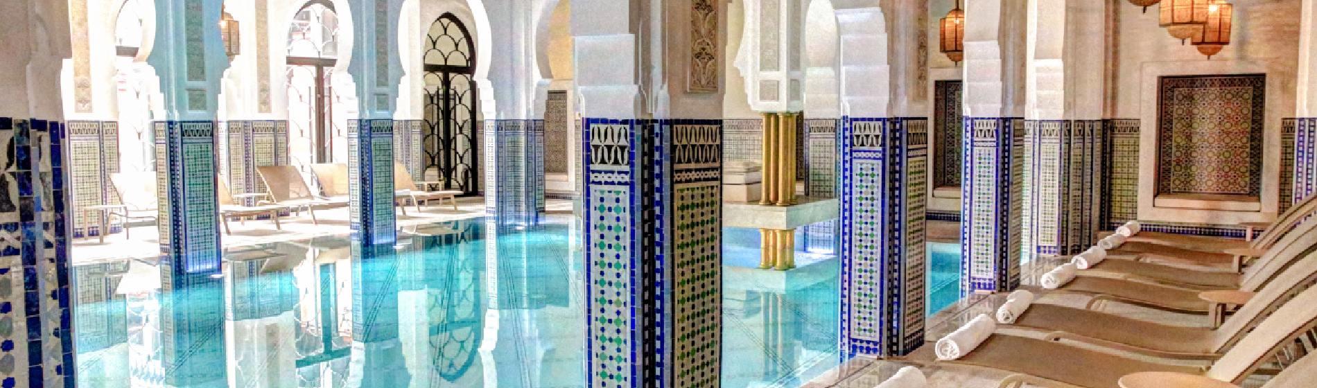 Indoor spa pool in a Moroccan-style resort with arched doorways, intricate mosaic tiles, turquoise water and cushioned loungers arranged beside the pool.