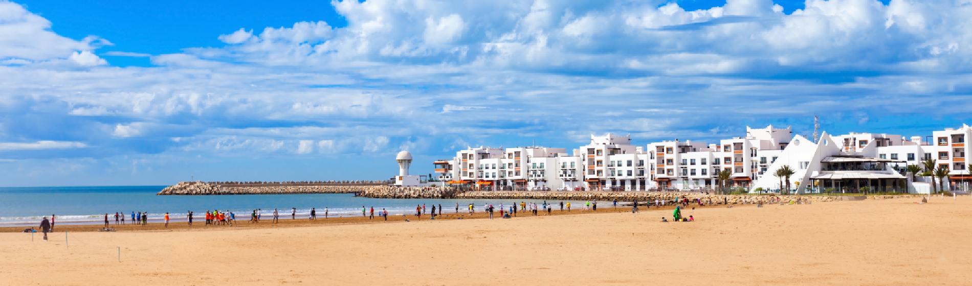 Sandy beach in Agadir with people walking along the shore, calm Atlantic water, and white resort buildings lining the promenade under a bright blue sky with scattered clouds.