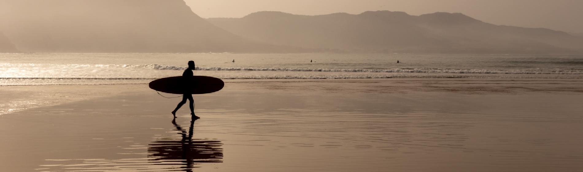 Silhouette of a surfer walking along Taghazout beach at sunset with waves and mountains in the background.