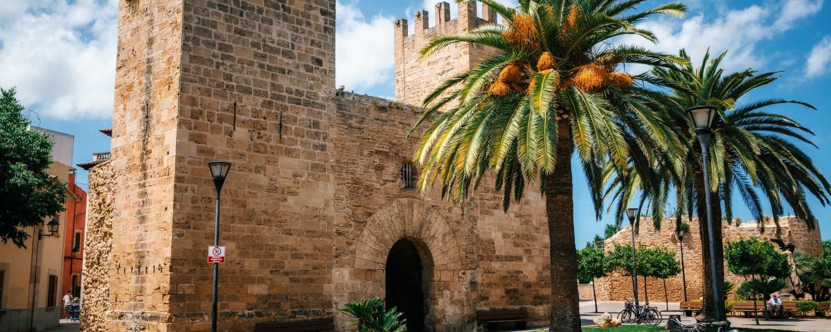 Imposing stone gatehouse from the medieval walls of Alcudia's Old Town, with buildings and someone on a bench in the background, a blue sky above, and palm trees in the foreground.