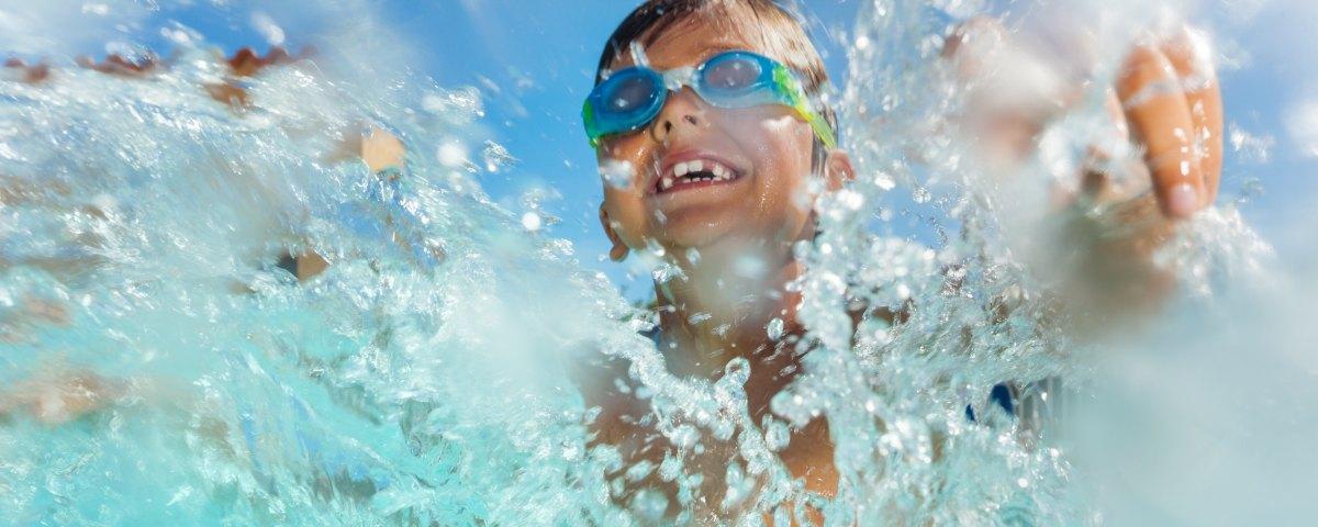 Close up of a smiling boy in swimming goggles splashing water vigorously