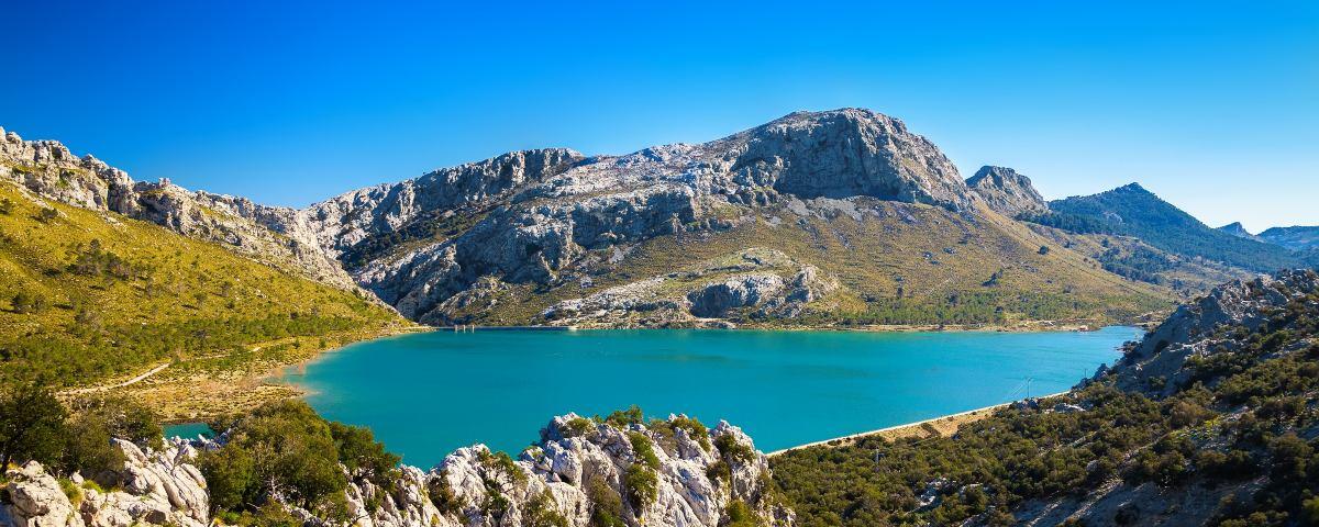 Turquoise blue lake surrounded by rocky outcrops and green vegetation, all under a clear blue sky in the Tramuntana Mountains of Majorca. There are mountain peaks in the background.