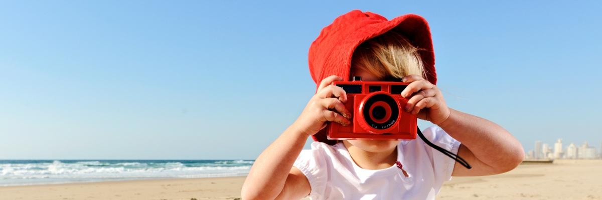 Small child in a white tee shirt and red hat on a white sand beach, taking photos with a red camera. The sky is clear and blue, and the sea is lapping the sand in the background.