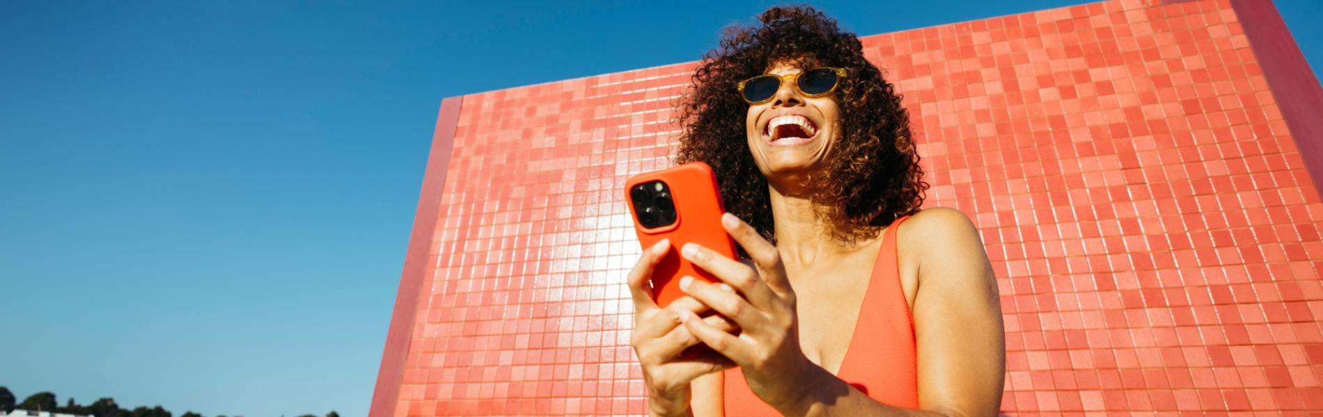 Smiling woman in sunglasses using her phone against a red tiled wall and blue sky.