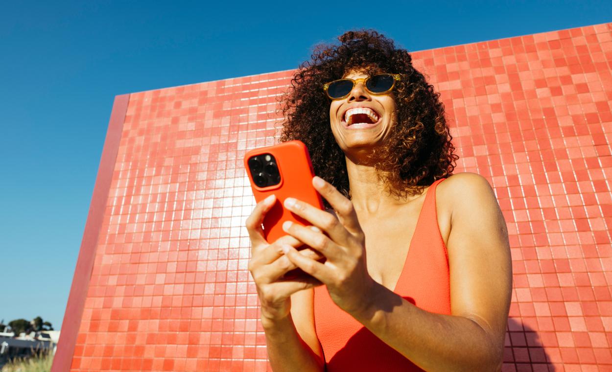 Smiling woman in sunglasses using her phone against a red tiled wall and blue sky.