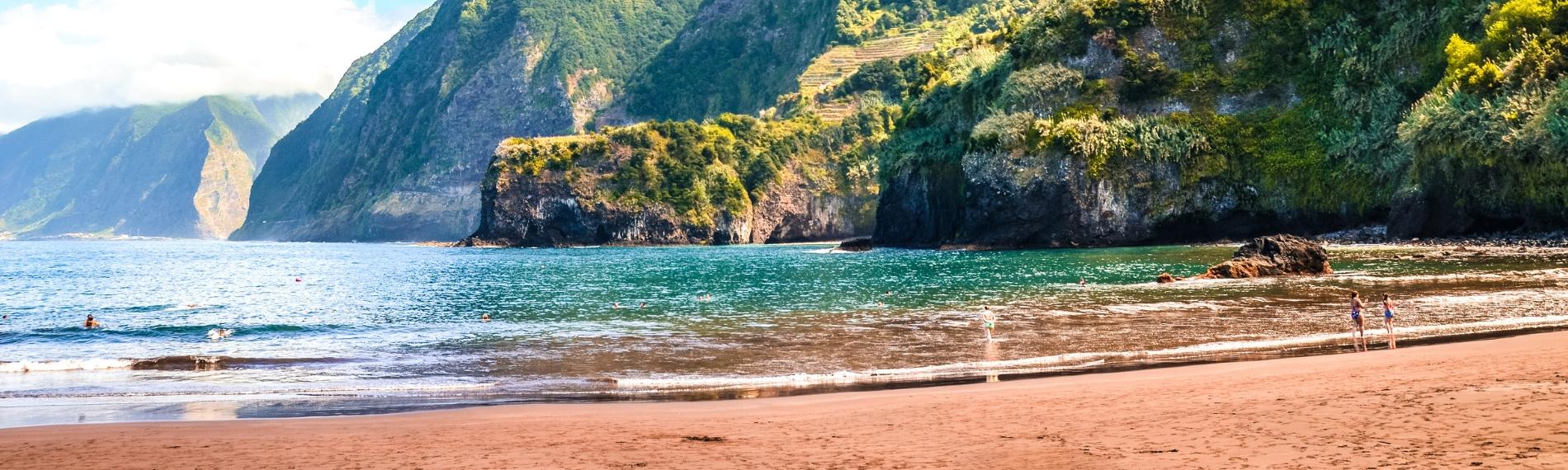 People swimming at a sandy beach beneath steep, green mountains in Madeira.