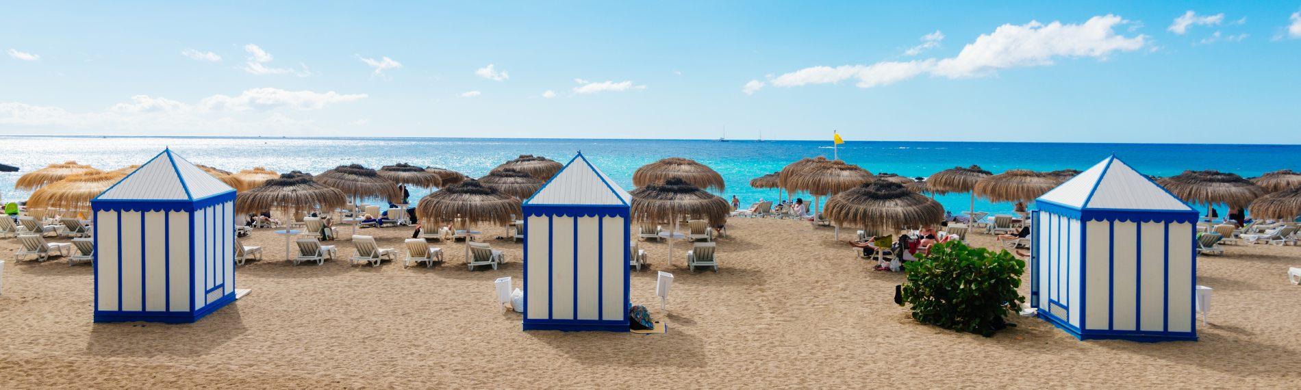 Looking out to the blue sea, past stripey huts and thatched parasols, on the sandy beach of Playa del Duque in Tenerife