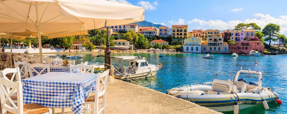 Restaurant tables on the waterfront in Assos, Kefalonia, with a view of the harbour, boats and colourful buildings typical of the island. The tables have blue and white checked tablecloths, cream parasols, and white wooden chairs.