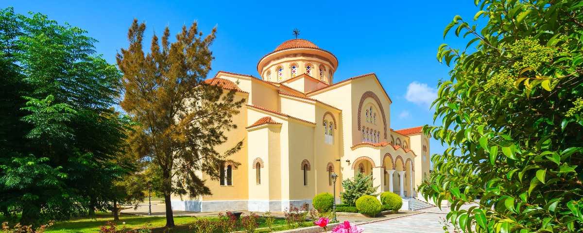 Grand-looking building with creamy walls, terracotta roof tiles and arched doors and windows surrounded by a neat garden and driveway. This is part of the Monastery of Agios Gerasimos, a fascinating place to visit in Kefalonia, Greece.