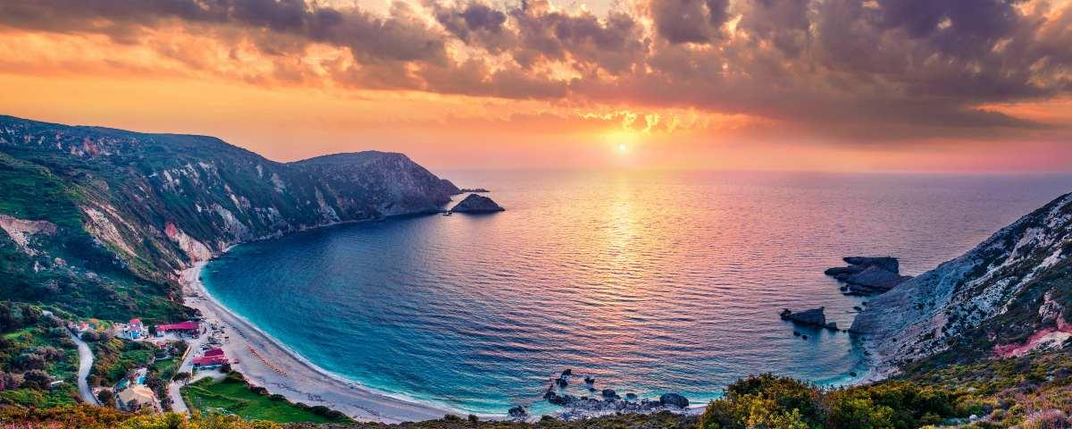 Golden sunset over the Ionian Sea and Petani Beach in Kefalonia. The beach is in a bay wrapped with gentle cliffs. We're looking across the beach and bay towards the horizon, where the sun is low in the sky, below the evening clouds, lighting up the sky yellow and reflecting in the calm sea.