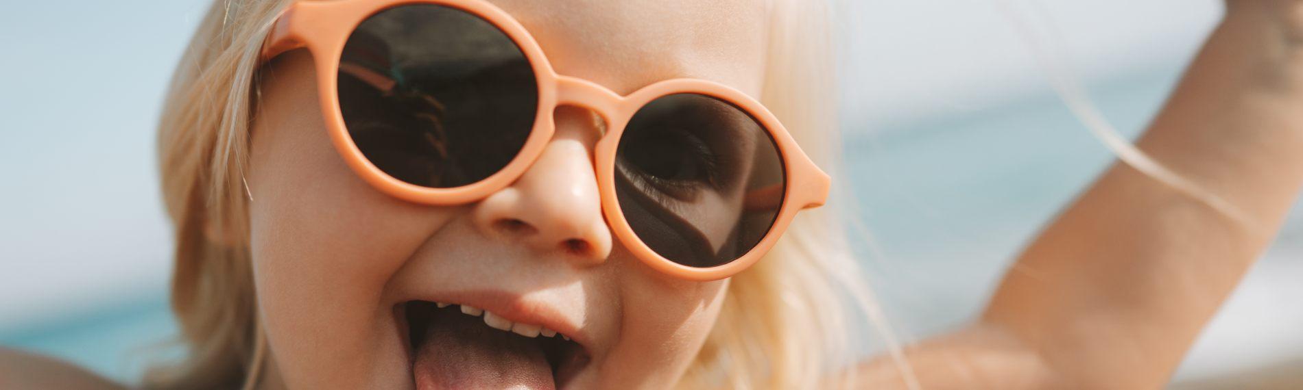 Happy toddler on a beach, wearing peach-framed sunglasses, sticking tongue out and waving arms in the air