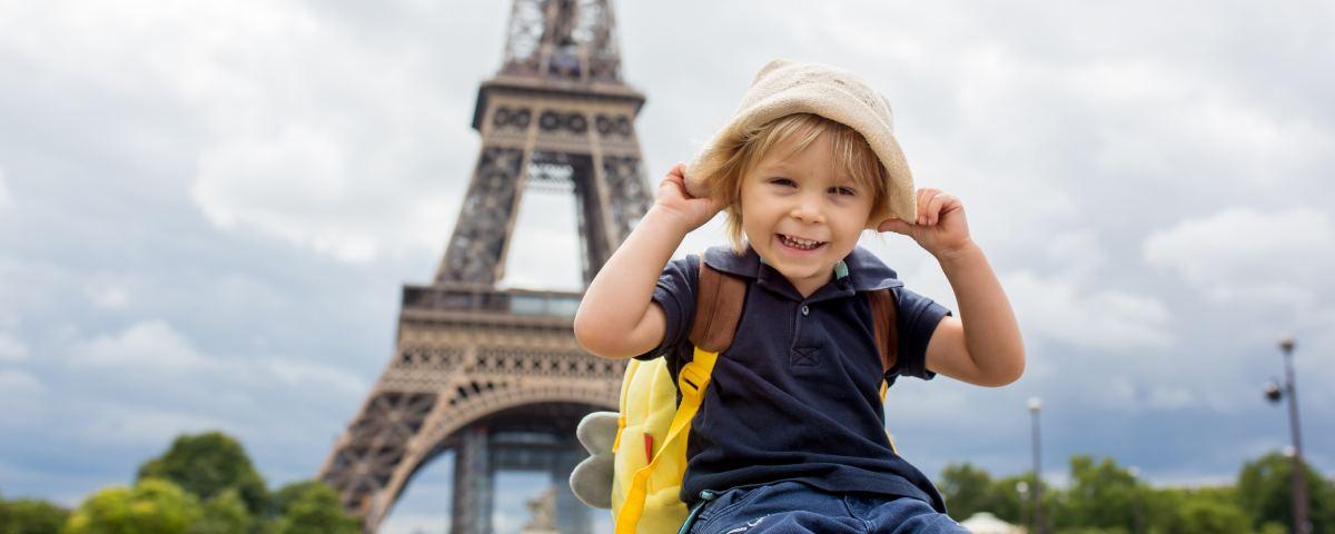 Smiling young child sitting in front of the Eiffel Tower, wearing a sun hat and a yellow rucksack.
