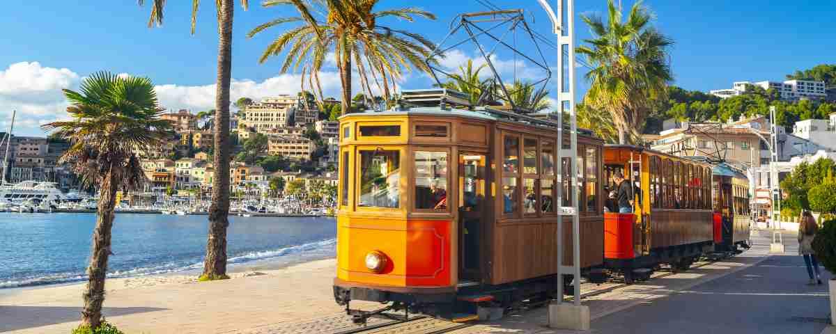 The historic orange tram that runs between Soller and Port de Soller in Mallorca, here shown on the seafront with palm trees and a sunny blue sky with boats in the background.
