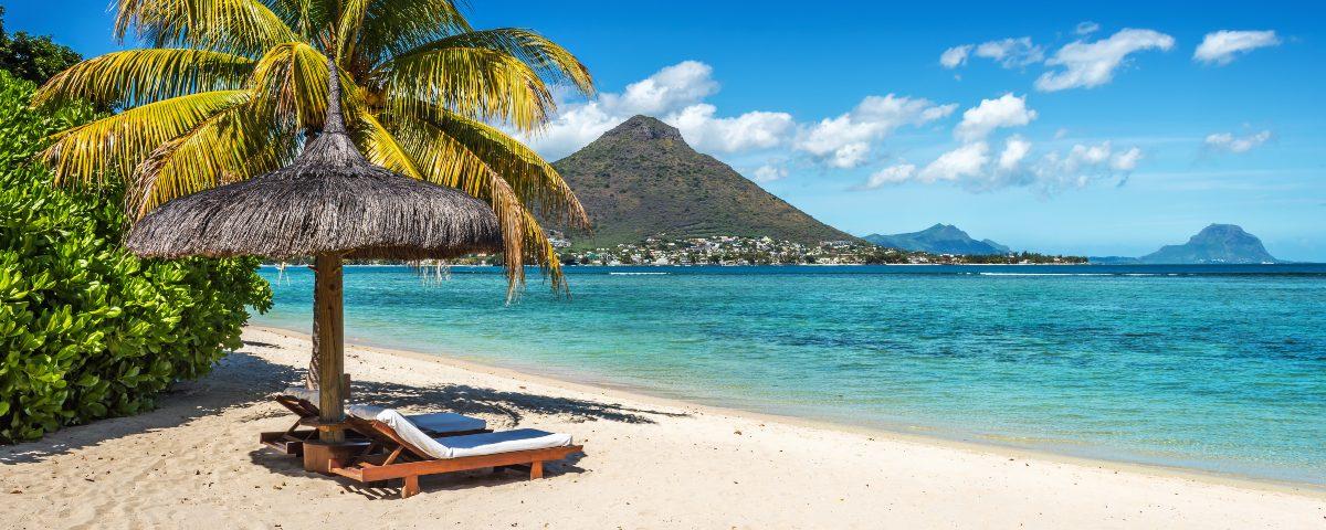 Two sunloungers under a thatched parasol on a white-sand beach in Mauritius, with a view of a mountain