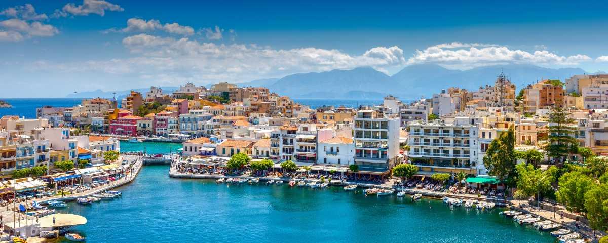 View of Agios Nikolaos harbour in Crete, with blue water and colourful buildings of all shapes and sizes. Glimpse of the sea and shadowy hills in the background.