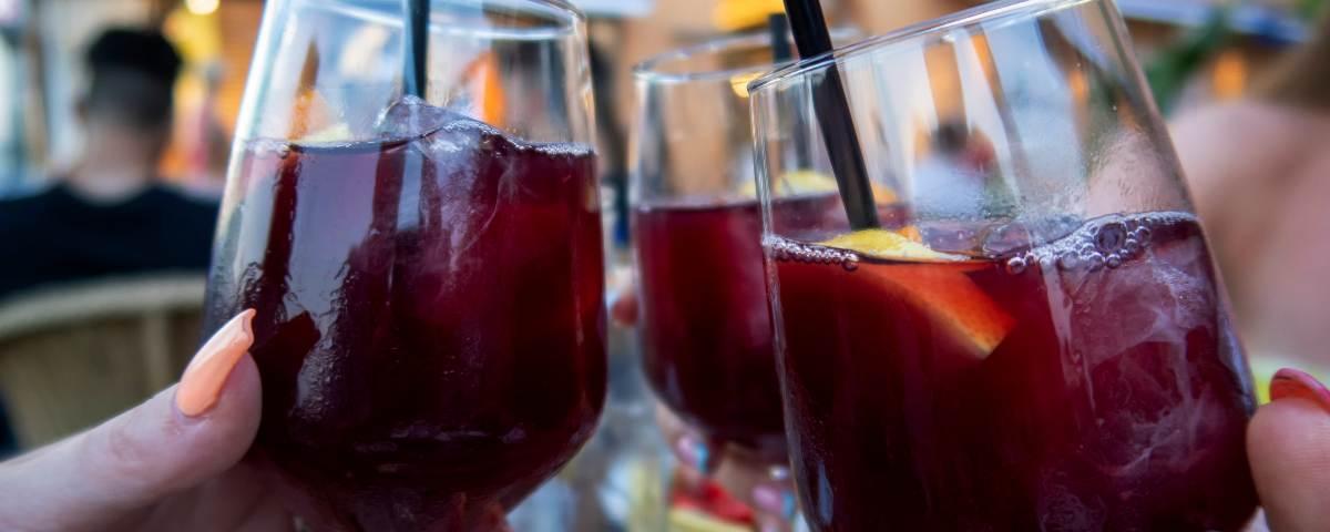 Three glasses of sangria with black straws and floating fruit, held up in cheers position. People sitting at al fresco tables can be seen out of focus in the background