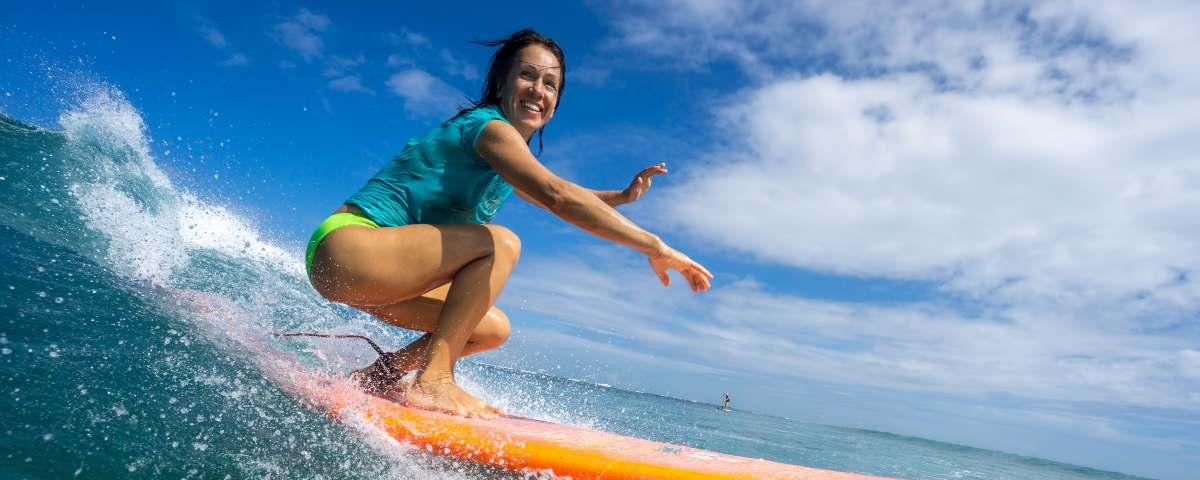 Smiling woman on an orange board and wearing a turquoise tee shirt and fluorescent bikini bottoms, looking at the camera and surfingf on the Portuguese coast