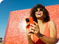 Smiling woman in sunglasses using her phone against a red tiled wall and blue sky.